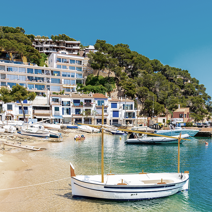 Puerto y playa de Llafranc con barcas sobre la arena, una menorquina y viviendas junto al puerto deportivo.